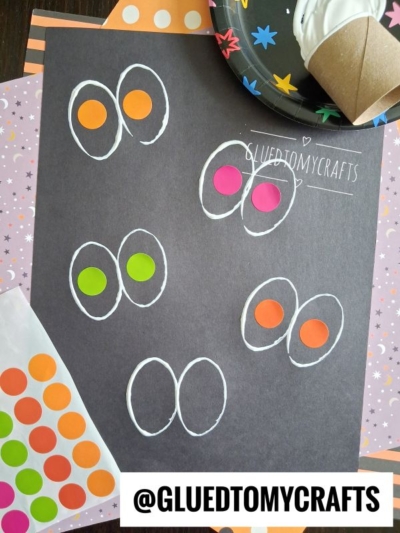 A black paper with hand-drawn white ovals, each pair filled with colorful round stickers, creating eye shapes—perfect for Halloween Crafts for Kids. Craft supplies and a plate with a cup are nearby. The text @GLUEDTOMYCRAFTS is shown on the paper.