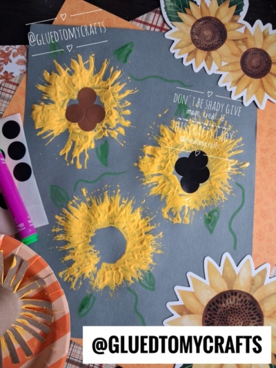 A gray paper displays three painted sunflower crafts, featuring green stems, paper petals, and brown circle centers—perfect examples of FALL CRAFTS FOR KIDS. Art supplies, flower cutouts, and a purple marker are scattered around the paper. @GLUEDTOMYCRAFTS is written on the image.