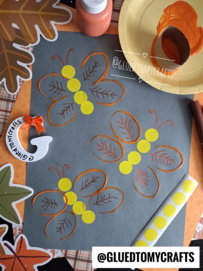 A fall-themed craft setup perfect for FALL CRAFTS FOR KIDS, featuring three butterfly prints made from a painted toilet paper roll, yellow sticker dots for bodies, and leaf decorations scattered around the workspace. Supplies like paint, brush, and paper are also visible.