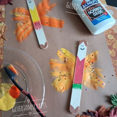 Two popsicle stick fairies with painted wings on brown paper, surrounded by fake leaves, a toothbrush with paint, and a bottle of Elmers glue. The popsicle sticks have smiling faces and colorful bodies.