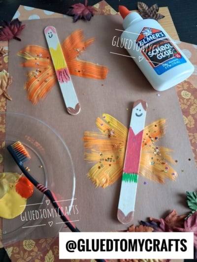 Two popsicle stick fairies with painted wings on brown paper, surrounded by fake leaves, a toothbrush with paint, and a bottle of Elmers glue. The popsicle sticks have smiling faces and colorful bodies.
