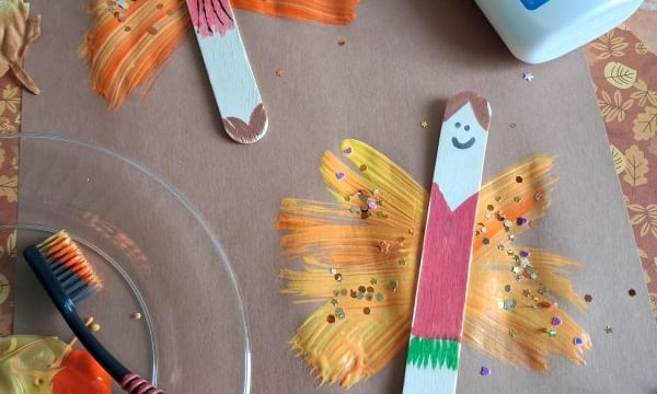 Two popsicle stick fairies with painted wings on brown paper, surrounded by fake leaves, a toothbrush with paint, and a bottle of Elmers glue. The popsicle sticks have smiling faces and colorful bodies.
