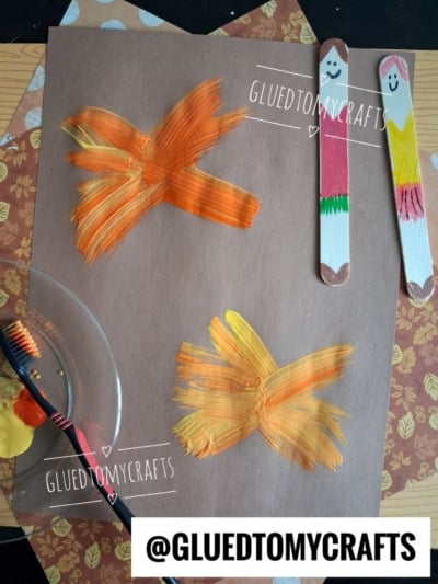 Brown paper with two orange-yellow painted X shapes, two decorated craft sticks with faces and clothes, a paintbrush beside a plate of yellow paint, and GLUEDTOMYCRAFTS watermarks on the image.