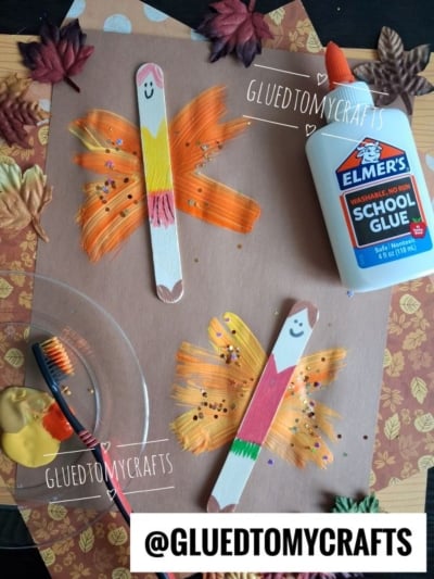 Two popsicle stick crafts with painted wings, glitter, and smiley faces on brown paper. Elmers glue, a paintbrush, yellow paint, and autumn leaves are nearby. @GLUEDTOMYCRAFTS is labeled on the image.