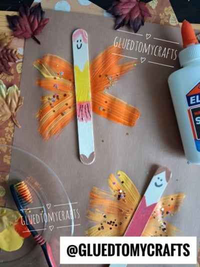 Two butterfly crafts made from painted popsicle sticks and colorful paint on brown paper, with glue, leaves, glitter, and a paintbrush nearby. The popsicle sticks have smiling faces drawn on them.