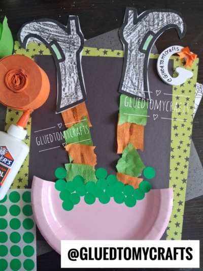 Craft project featuring crepe paper witch legs made from paper, green dots, and orange tissue, sticking out of a pink paper plate. Various craft supplies, spooky-themed papers, and an @GLUEDTOMYCRAFTS tag are visible.