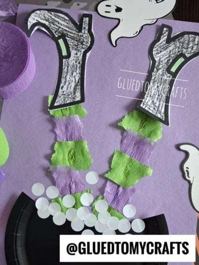 A Halloween craft shows a witchs legs made of torn green and purple paper, black boots, and white circles as bubbles in a black cauldron on purple paper. Ghost and crepe paper shapes decorate the background.