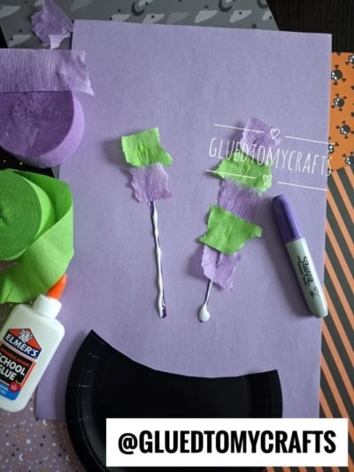 Purple paper with glue lines, torn green and purple crepe paper, Elmers glue, a purple marker, and black plates. Craft materials are arranged for a project. @GLUEDTOMYCRAFTS is labeled at the bottom.
