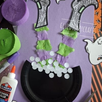 A Halloween craft shows a witchs legs made of paper and streamers sticking out of a black cauldron, surrounded by ghosts, glue, and rolls of green and purple crepe paper.