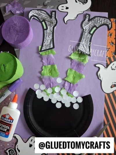 A Halloween craft shows a witchs legs made of paper and streamers sticking out of a black cauldron, surrounded by ghosts, glue, and rolls of green and purple crepe paper.