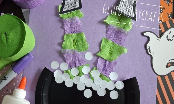 A Halloween craft shows a witchs legs made of paper and streamers sticking out of a black cauldron, surrounded by ghosts, glue, and rolls of green and purple crepe paper.