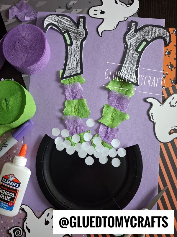A Halloween craft shows a witchs legs made of paper and streamers sticking out of a black cauldron, surrounded by ghosts, glue, and rolls of green and purple crepe paper.