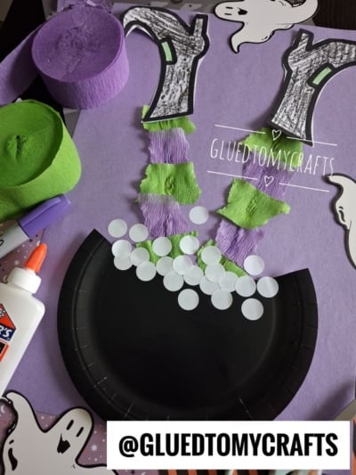 Halloween witch legs craft with green and purple crepe paper, black paper plate, paper ghosts, glue bottle, and @GLUETOMYCRAFTS label on purple background. Art supplies are scattered around the crafting area.