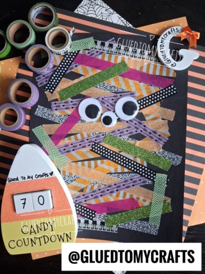 A delightful Halloween craft project perfect for Halloween Crafts for Kids, this activity features a mummy made from colored tape strips on black paper, complete with googly eyes, washi tape rolls, and a candy countdown card set against a striped orange and black background.