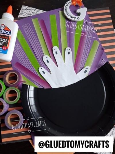 Halloween craft supplies: black plate, paper hand cutout with ghost faces, glue, washi tape, and colored paper arranged on a surface. @GLUEDTOMYCRAFTS watermark appears on the image.