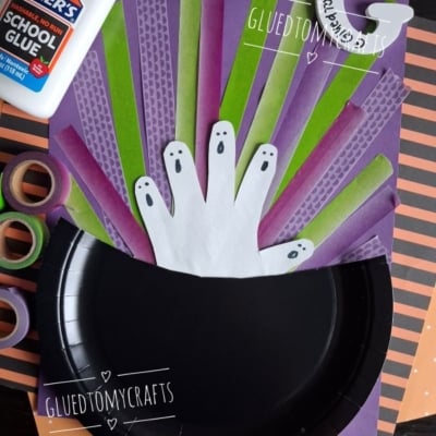 A Halloween craft shows paper ghost hands in a black paper cauldron with green and purple strips, Elmers glue, washi tape, and an orange-striped background. The username @GLUEDTOMYCRAFTS is displayed.