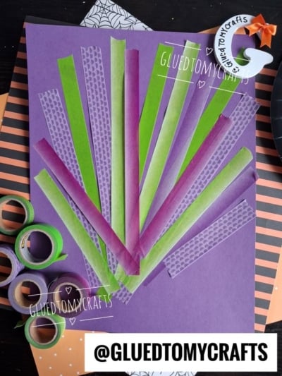 A purple paper with green and purple washi tape strips arranged in a fan shape. Rolls of tape are nearby, along with Halloween-themed decorations. @GluedToMyCrafts is labeled on the image.