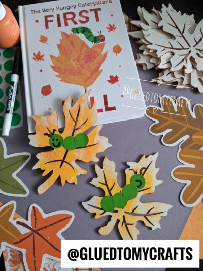 Children’s fall craft project: Paper leaves decorated with green paper caterpillars—perfect for FALL CRAFTS FOR KIDS. The book The Very Hungry Caterpillar’s First Fall is in the background, with craft supplies and cutout leaves scattered around.