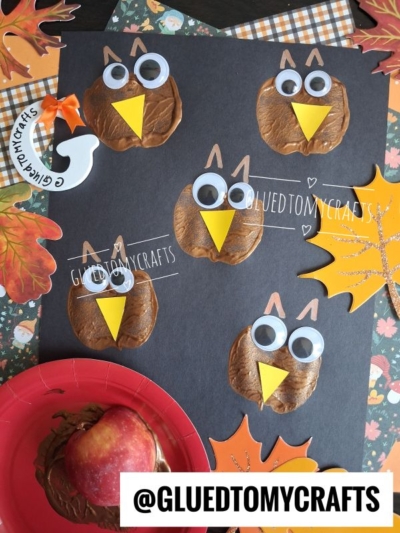 Five brown owl crafts with googly eyes and yellow beaks made from painted apple prints are displayed on black paper, surrounded by autumn leaves and seasonal decorations. Showcasing a creative apple stamped craft idea, a red plate with a painted apple is shown in the corner.
