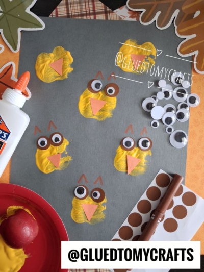 Childrens owl art project with yellow paint, brown and pink paper shapes, googly eyes, a bottle of glue, and autumn leaf decorations on a gray sheet. Supplies and finished owls are arranged on a tabletop, creating a festive scene perfect for an APPLE STAMPED CRAFT or other fun fall activities.
