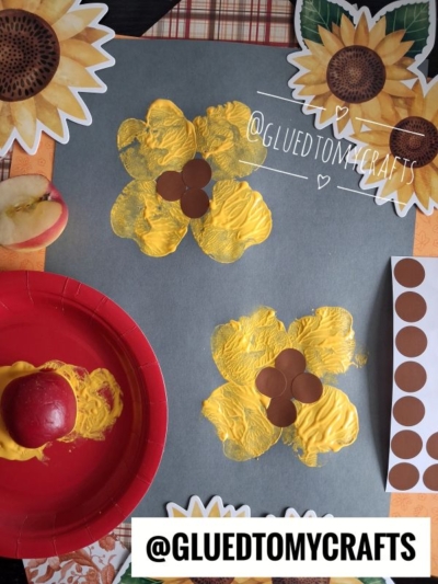 A gray sheet with yellow sunflower stamps, brown dot stickers, a paint-covered apple, and sunflower decorations; art supplies arranged on a table for an easy apple stamped craft.