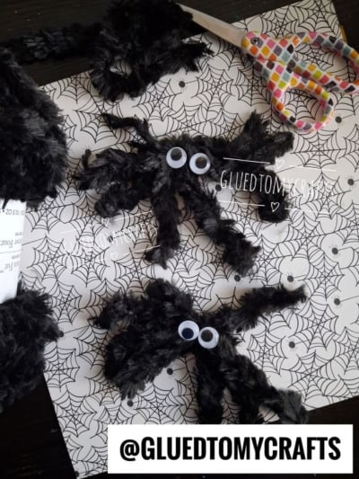 Two fuzzy black spiders with googly eyes sit on spiderweb-patterned paper, next to a pair of scissors with colored handles—making this scene perfect inspiration for Halloween Crafts for Kids. The text @GLUEDTOMYCRAFTS appears at the bottom.