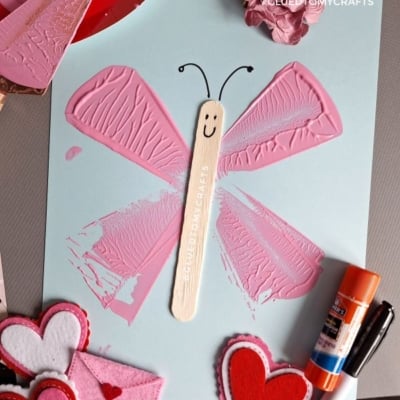 A butterfly craft made with pink painted wings, a craft stick body with a smiley face, and black antennae, displayed with felt hearts, a glue stick, and pink paper flowers. @GLUEDTOMYCRAFTS text at the bottom.