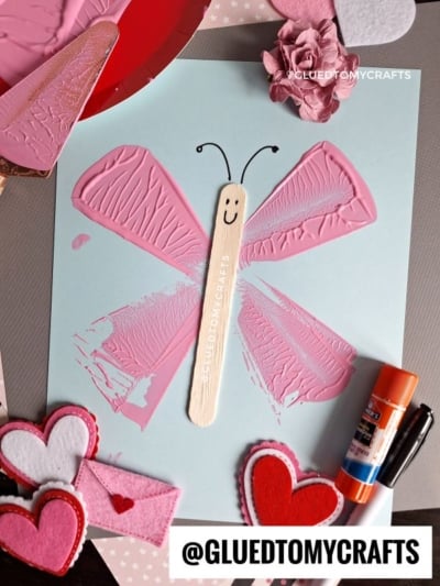 A butterfly craft made with pink painted wings, a craft stick body with a smiley face, and black antennae, displayed with felt hearts, a glue stick, and pink paper flowers. @GLUEDTOMYCRAFTS text at the bottom.