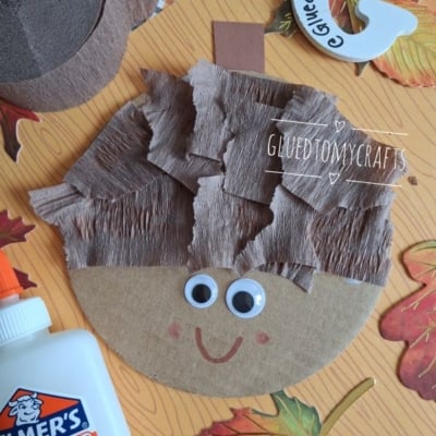 A cardboard circle with googly eyes, a red smile, and brown crepe paper hair, resembling a childs craft face. School glue, leaves, and craft materials are around it. @GLUEDTOMYCRAFTS text is shown.