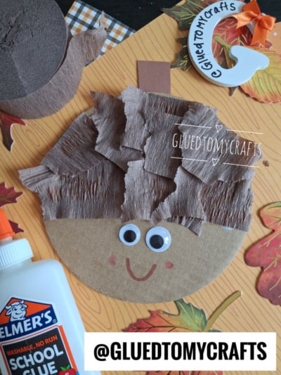 A cardboard circle with googly eyes, a red smile, and brown crepe paper hair, resembling a childs craft face. School glue, leaves, and craft materials are around it. @GLUEDTOMYCRAFTS text is shown.