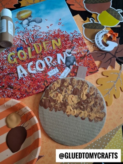 A fall-themed craft setup with a book titled The Golden Acorn, paint, brushes, paper acorns, and a painted cardboard acorn on a table decorated with autumn-colored paper leaves—perfect inspiration for FALL CRAFTS FOR KIDS. @GLUEDTOMYCRAFTS is written at the bottom.