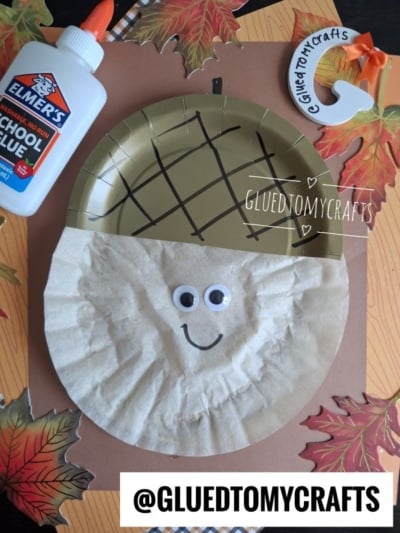 A coffee filter acorn craft with googly eyes and a smiling face, created using a gold plate, brown liner, and glue, surrounded by autumn leaves and @GLUEDTOMYCRAFTS tags.
