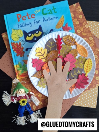 Childrens autumn craft with a paper plate, paper hand, felt leaves, and glitter. Pete the Cat book, fall-themed scrapbook paper, and a small scarecrow also shown. @GLUEDTOMYCRAFTS watermark at the bottom.