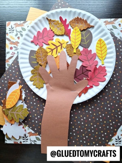 A paper hand is glued to a plate and surrounded by colorful paper leaves on polka dot paper. The crafting materials are fall-themed. The text @GLUEDTOMYCRAFTS is at the bottom.