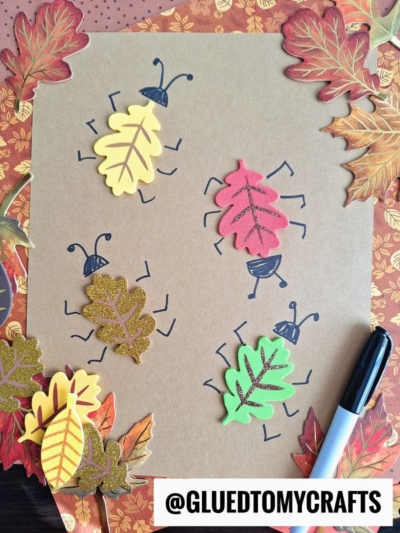 Four colorful foam leaves with drawn legs and antennae resemble bugs on brown paper, surrounded by autumn leaves. A black marker and @GLUEDTOMYCRAFTS label appear at the bottom right.