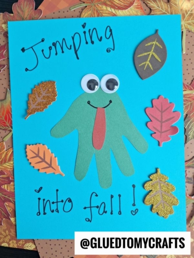 A blue card with a green handprint frog, googly eyes, and red tongue, surrounded by colorful fall leaf stickers. Text reads Jumping into fall! Card sits on a fall leaf background.