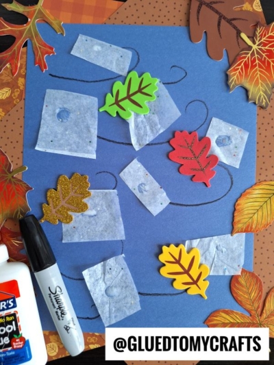 Blue paper with wavy lines, square tissue pieces, colorful foam leaves, glue, a Sharpie, and autumn leaf decorations on the sides. @GLUEDTOMYCRAFTS is labeled at the bottom.