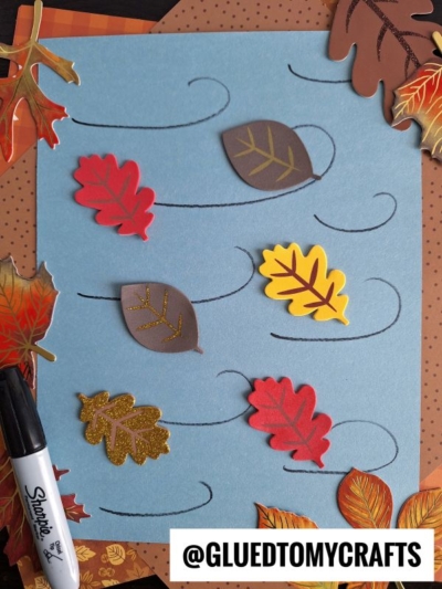 A blue paper with swirly black lines and colorful paper leaves scattered on it, surrounded by autumn-themed decorations and a Sharpie marker. The handle @GLUEDTOMYCRAFTS is in the bottom right corner.
