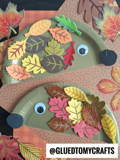 Two paper plate hedgehog crafts decorated with colorful paper and foam leaves, googly eyes, and placed on dotted paper with autumn leaves scattered around. A @GLUEDTOMYCRAFTS label is at the bottom.