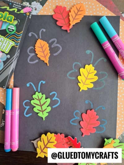 Black paper with colorful leaf stickers arranged like butterflies, surrounded by bright markers and craft supplies. The text @GLUEDTOMYCRAFTS is at the bottom on a white label.