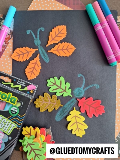 Colorful leaf-shaped cutouts arranged as butterfly wings on black paper, with teal crayon bodies drawn in the center. Markers and a Crayola pack are nearby. @GLUEDTOMYCRAFTS is in bold at the bottom.
