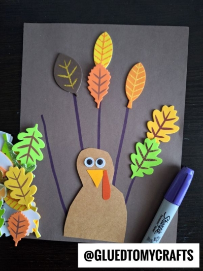 A turkey craft made with brown paper, googly eyes, and colorful foam leaves for feathers on a dark background. A purple marker and extra foam leaves are beside it. @GLUEDTOMYCRAFTS appears in the corner.