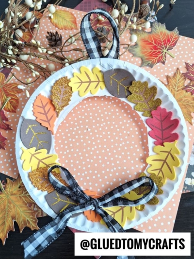 A paper plate fall wreath decorated with colorful paper leaves, a black-and-white plaid ribbon bow, and set on a polka-dot background with autumn leaves and berries. @GLUETOMYCRAFTS appears at the bottom.