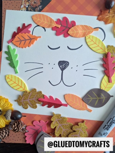 A paper with a simple lion face drawn on it, surrounded by colorful foam and paper leaves. Autumn-themed craft supplies are scattered around, with a @GLUEDTOMYCRAFTS label at the bottom.