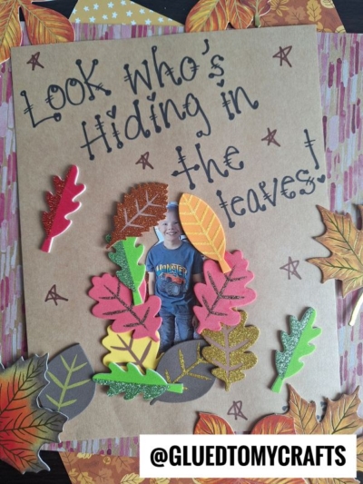 A craft page with colorful paper leaves hiding a childs photo, handwritten text reads Look whos hiding in the leaves! Autumn leaves decorate the background; @GLUEDTOMYCRAFTS is written at the bottom.