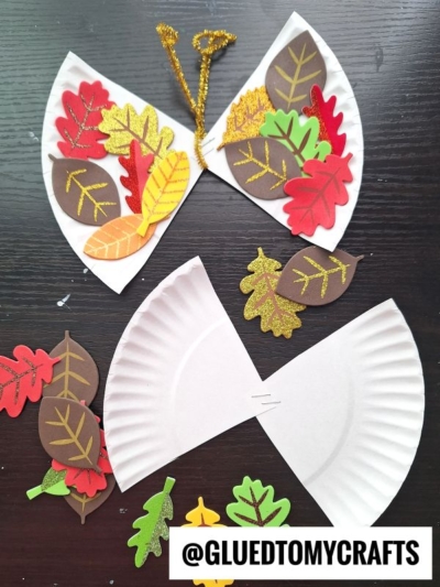 A butterfly craft made from a cut paper plate, decorated with colorful felt and foam leaves. Loose leaves and crafting supplies are scattered nearby. The text @GLUEDTOMYCRAFTS is at the bottom.