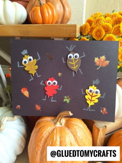 A black paper with leaf characters, googly eyes, and arms and legs made from paper, surrounded by fall leaf stickers. Pumpkins and gourds are in the background. The handle @GLUEDTOMYCRAFTS is at the bottom.