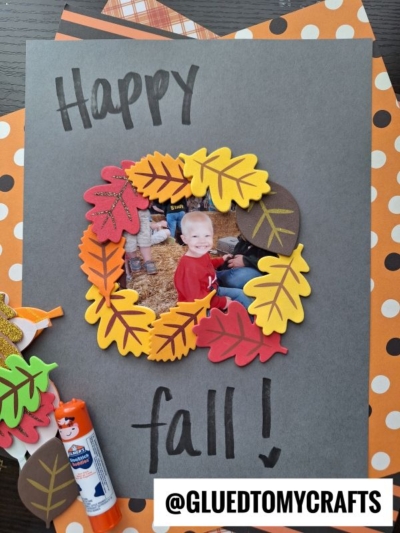 A fall-themed craft features a photo of a smiling child framed by colorful paper leaves on black paper, with Happy fall! handwritten above and below. Craft supplies and patterned papers are nearby.