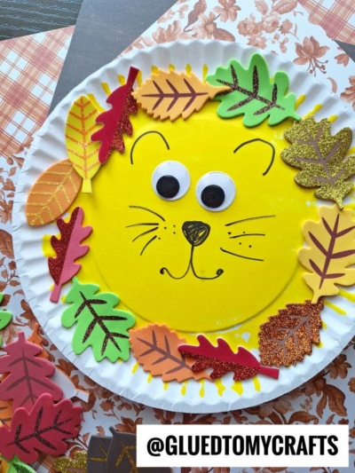 A paper plate craft of a lions face with googly eyes, drawn nose, mouth, and whiskers, surrounded by colorful, decorative autumn leaves.