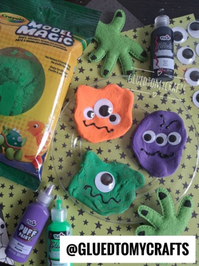 Colorful clay monsters with googly eyes on a star-patterned surface, surrounded by green felt shapes, paint bottles, and a Crayola Model Magic package—perfect for fun Halloween Crafts for Kids. @GLUEDTOMYCRAFTS is written on the image.